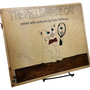 Può includere: Libro per bambini vintage intitolato "THE PROUD WHITE CAT" con un'illustrazione a cartoni animati di un gatto bianco che si guarda allo specchio. Il libro ha una copertina beige con testo nero e un supporto in metallo nero.
