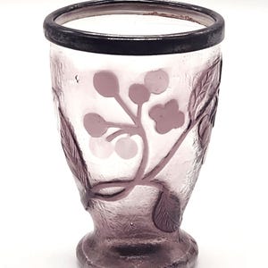 May include: A vintage, amethyst-colored glass vase with a floral design. The vase features a dark rim and a detailed pattern of leaves and berries. The design is etched into the glass, creating a textured surface. The vase has a rounded base and a flared top.