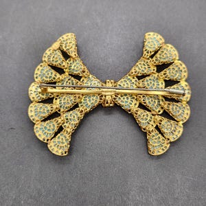 Peut inclure: Broche dor&eacute;e en forme de n&oelig;ud papillon, avec un travail ajour&eacute; complexe et un fond bleu clair. La broche est dot&eacute;e d'une fermeture &agrave; &eacute;pingle au dos. Le design est sym&eacute;trique, avec un motif d&eacute;taill&eacute; et dentel&eacute;. La broche est probablement vintage ou antique.