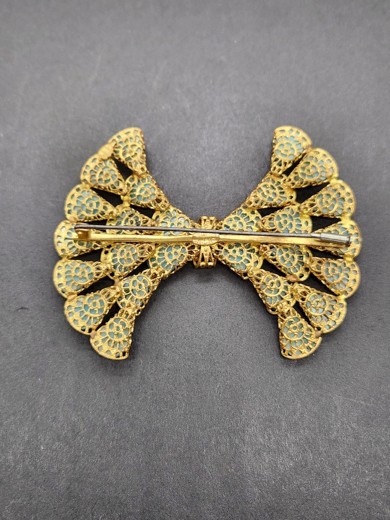 Peut inclure: Broche dor&eacute;e en forme de n&oelig;ud papillon, avec un travail ajour&eacute; complexe et un fond bleu clair. La broche est dot&eacute;e d'une fermeture &agrave; &eacute;pingle au dos. Le design est sym&eacute;trique, avec un motif d&eacute;taill&eacute; et dentel&eacute;. La broche est probablement vintage ou antique.
