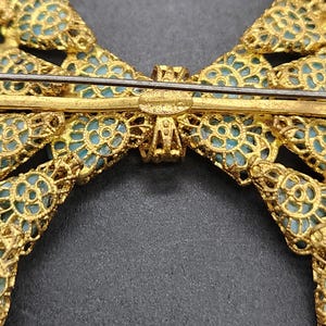 Peut inclure: Broche en forme de n&oelig;ud papillon dor&eacute;, avec un travail ajour&eacute; complexe et un fond bleu clair. La broche porte une &eacute;pingle de fermeture et l'inscription "MADE IN JAPAN". Le design du n&oelig;ud est sym&eacute;trique et d&eacute;taill&eacute;.