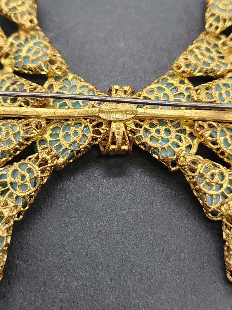 Peut inclure: Broche en forme de n&oelig;ud papillon dor&eacute;, avec un travail ajour&eacute; complexe et un fond bleu clair. La broche porte une &eacute;pingle de fermeture et l'inscription "MADE IN JAPAN". Le design du n&oelig;ud est sym&eacute;trique et d&eacute;taill&eacute;.