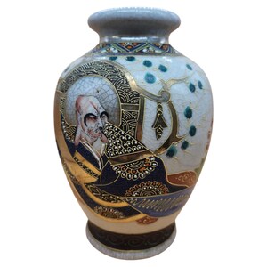 May include: An antique Japanese vase with a detailed illustration of a bald man in traditional attire. The vase features a light blue and gold colour scheme with intricate patterns and designs. The vase has a grey rim and base.