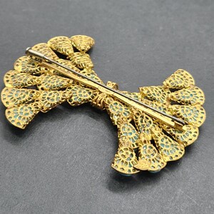 Peut inclure: Broche dor&eacute;e avec un design en filigrane complexe. La broche pr&eacute;sente une forme en &eacute;ventail avec des accents de couleur turquoise. L'&eacute;pingle est visible au dos, indiquant qu'elle peut &ecirc;tre fix&eacute;e aux v&ecirc;tements ou aux accessoires.