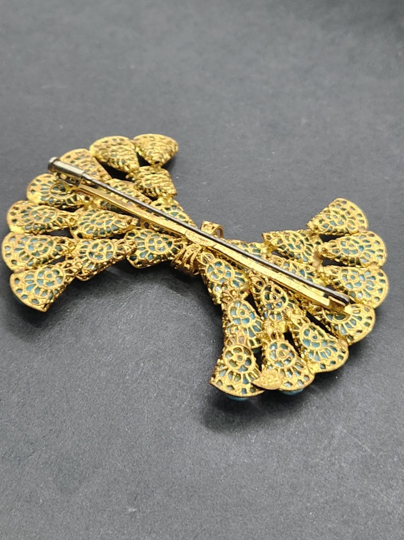 Peut inclure: Broche dor&eacute;e avec un design en filigrane complexe. La broche pr&eacute;sente une forme en &eacute;ventail avec des accents de couleur turquoise. L'&eacute;pingle est visible au dos, indiquant qu'elle peut &ecirc;tre fix&eacute;e aux v&ecirc;tements ou aux accessoires.