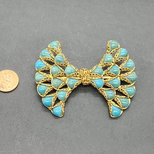 Peut inclure: Broche vintage en forme de n&oelig;ud papillon, orn&eacute;e de pierres en forme de goutte d'eau turquoise serties dans un cadre filigran&eacute; dor&eacute;. La broche mesure environ 7,6 cm de large, pos&eacute;e sur une surface gris fonc&eacute;.