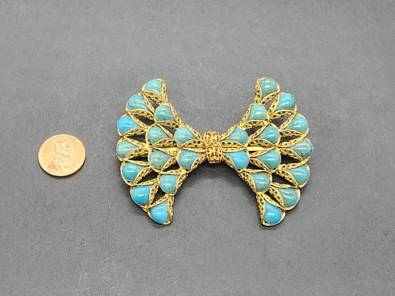 Peut inclure: Broche vintage en forme de n&oelig;ud papillon, orn&eacute;e de pierres en forme de goutte d'eau turquoise serties dans un cadre filigran&eacute; dor&eacute;. La broche mesure environ 7,6 cm de large, pos&eacute;e sur une surface gris fonc&eacute;.