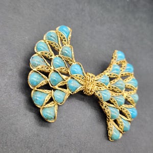 Peut inclure: Broche dor&eacute;e en forme de n&oelig;ud orn&eacute;e de pierres en forme de goutte d'eau de couleur turquoise. La broche pr&eacute;sente un design ajour&eacute; et un aspect textur&eacute;. Les pierres turquoises sont dispos&eacute;es sym&eacute;triquement, cr&eacute;ant un accessoire d&eacute;coratif.