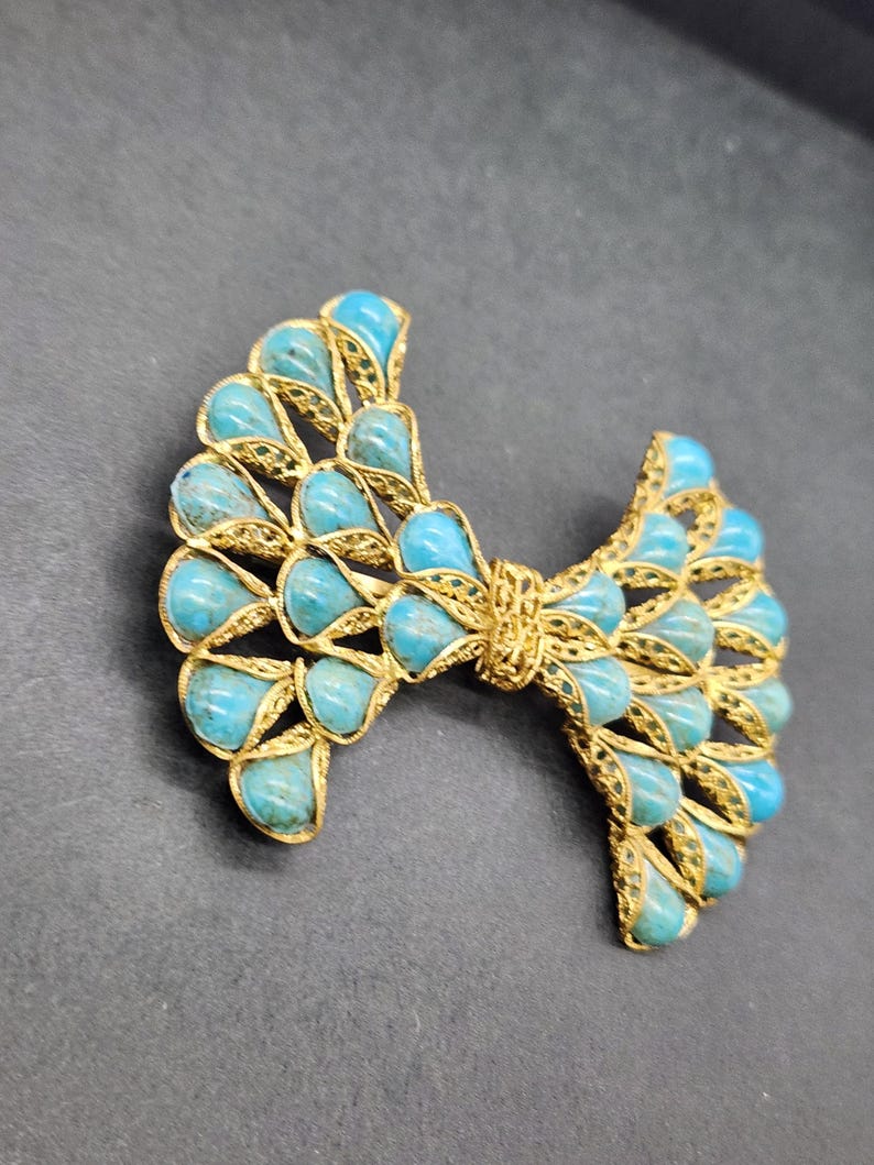 Peut inclure: Broche dor&eacute;e en forme de n&oelig;ud orn&eacute;e de pierres en forme de goutte d'eau de couleur turquoise. La broche pr&eacute;sente un design ajour&eacute; et un aspect textur&eacute;. Les pierres turquoises sont dispos&eacute;es sym&eacute;triquement, cr&eacute;ant un accessoire d&eacute;coratif.