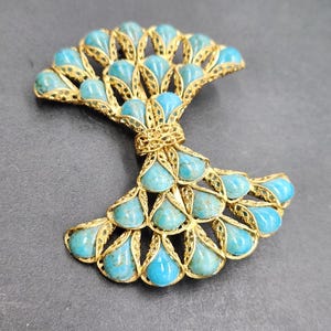 Peut inclure: Broche en m&eacute;tal dor&eacute; avec un motif en forme d'&eacute;ventail, orn&eacute;e de pierres en forme de goutte d'eau de couleur turquoise. La broche pr&eacute;sente un motif filigran&eacute; complexe, sur fond gris fonc&eacute;.