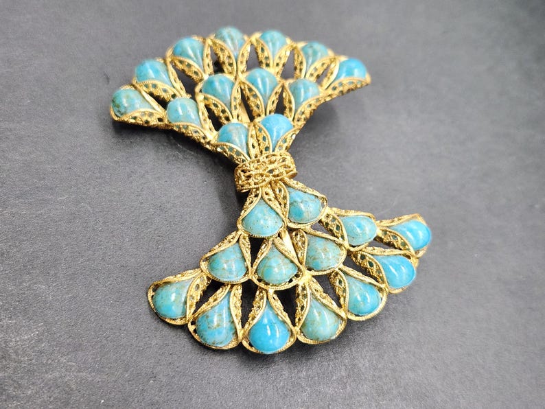 Peut inclure: Broche en m&eacute;tal dor&eacute; avec un motif en forme d'&eacute;ventail, orn&eacute;e de pierres en forme de goutte d'eau de couleur turquoise. La broche pr&eacute;sente un motif filigran&eacute; complexe, sur fond gris fonc&eacute;.