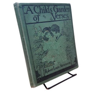 May include: A vintage book titled "A Child's Garden of Verses" by Robert Louis Stevenson. The cover is a faded teal green with black illustrations and text. The book is propped up on a black metal stand.