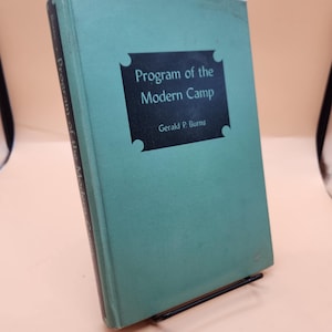 以下が含まれることがあります： ジェラルド・P・バーンズ著「Program of the Modern Camp」というタイトルの、ターコイズ色の本。タイトルは表紙の黒い長方形の中にあります。本は黒い金属製のスタンドに立てかけられています。