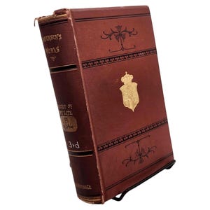 May include: A vintage, leather-bound book with the title "Andersen's Works" visible on the spine. The cover is a deep reddish-brown with gold embellishments, including a crest. The book is propped up on a black metal stand.