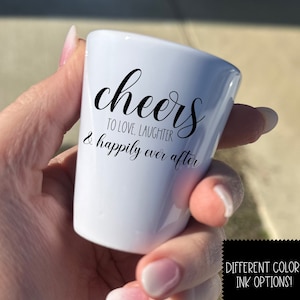 Puede incluir: Un vaso de chupito blanco con texto negro que dice "cheers to love, laughter & happily ever after".
