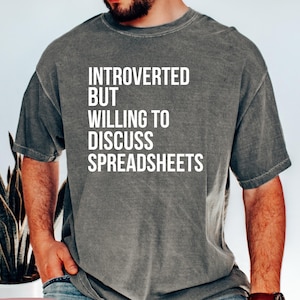 May include: A gray t-shirt with the text "INTROVERTED BUT WILLING TO DISCUSS SPREADSHEETS" in white. The shirt is a solid color and has short sleeves. The shirt is a Pepper color.