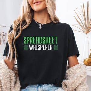 May include: Black t-shirt with the text "SPREADSHEET WHISPERER" in green and white. The shirt features a calculator graphic. The t-shirt is a comfortable, casual wear item.
