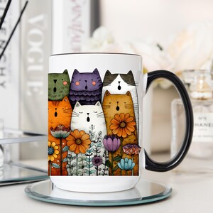 May include: White ceramic mug with a black handle featuring a colorful illustration of singing cats and flowers. The cats are in various colors, including orange, purple, and green. The flowers are orange, purple, and blue.