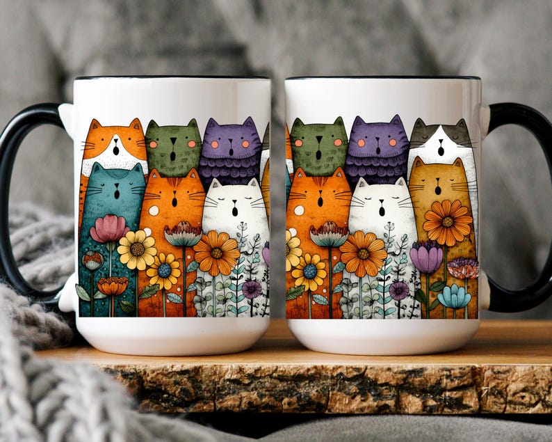 May include: Two white ceramic mugs with black handles and rims. Each mug features a colorful illustration of singing cats and flowers. The cats are in various colors, including orange, blue, and purple. The mugs are on a wooden surface.