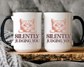 May include: Two white ceramic mugs with black handles and rims. Each mug features a peach-colored illustration of a cat and the text "SILENTLY JUDGING YOU" in a brown font. The mugs are on a wooden surface.