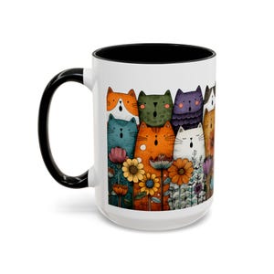 May include: White ceramic mug with a black handle and interior. The mug features a colorful illustration of singing cats with flowers. The cats are various colors, including orange, green, and blue.