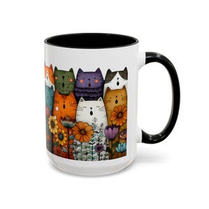 May include: White ceramic mug with a black interior and handle. The mug features a colorful illustration of cartoon cats and flowers. The cats are in various colors, including orange, green, and purple. The flowers are in shades of yellow, orange, and purple.