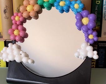 Puede incluir: Un espejo decorativo con base negra y marco floral colorido. Las flores son en tonos rosa, verde, azul y morado, con centros amarillos. Acentos blancos en forma de nube en la parte inferior.