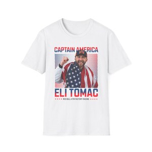 Peut inclure: T-shirt blanc avec un graphisme d'Eli Tomac, le texte "CAPTAIN AMERICA" au-dessus et "ELI TOMAC" en dessous. L'image comprend le drapeau am&eacute;ricain et le logo Red Bull KTM Factory Racing.