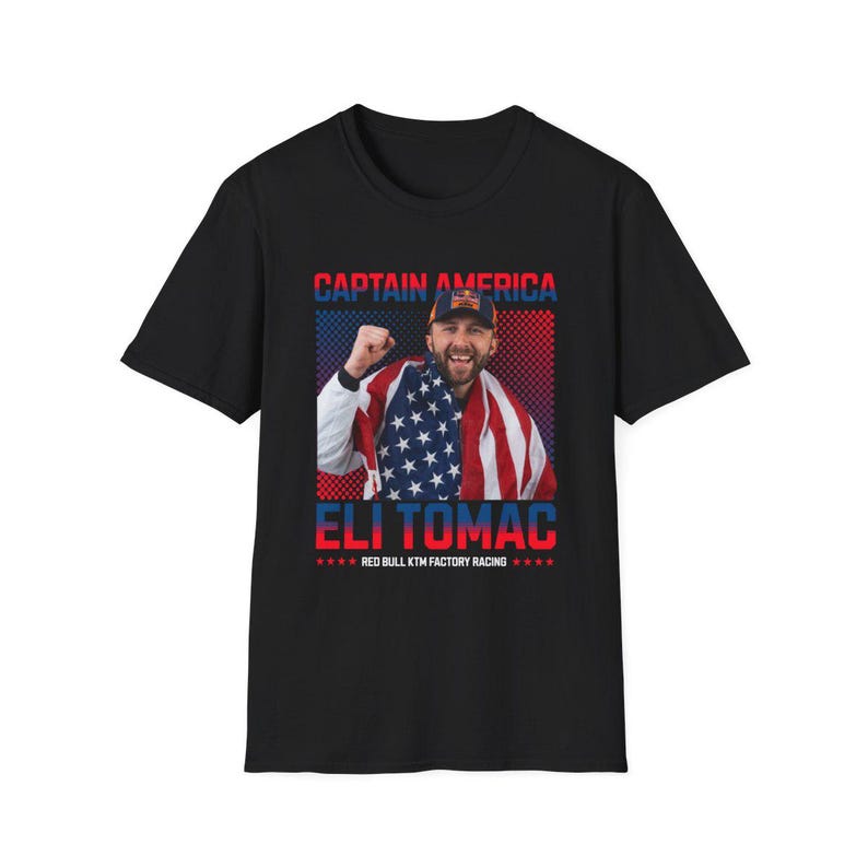Peut inclure: T-shirt noir avec un graphisme d'Eli Tomac, le texte "CAPTAIN AMERICA" au-dessus et "ELI TOMAC" en dessous. Le design comprend une image d'Eli Tomac avec un drapeau am&eacute;ricain. Le texte inf&eacute;rieur indique "RED BULL KTM FACTORY RACING".
