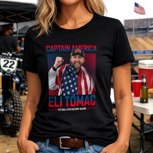 Peut inclure: T-shirt noir &agrave; col rond avec un graphisme d'un homme envelopp&eacute; dans un drapeau am&eacute;ricain. Le texte "CAPTAIN AMERICA" et "ELI TOMAC" sont affich&eacute;s au-dessus et en dessous de l'image, avec "RED BULL KTM FACTORY RACING" en dessous.