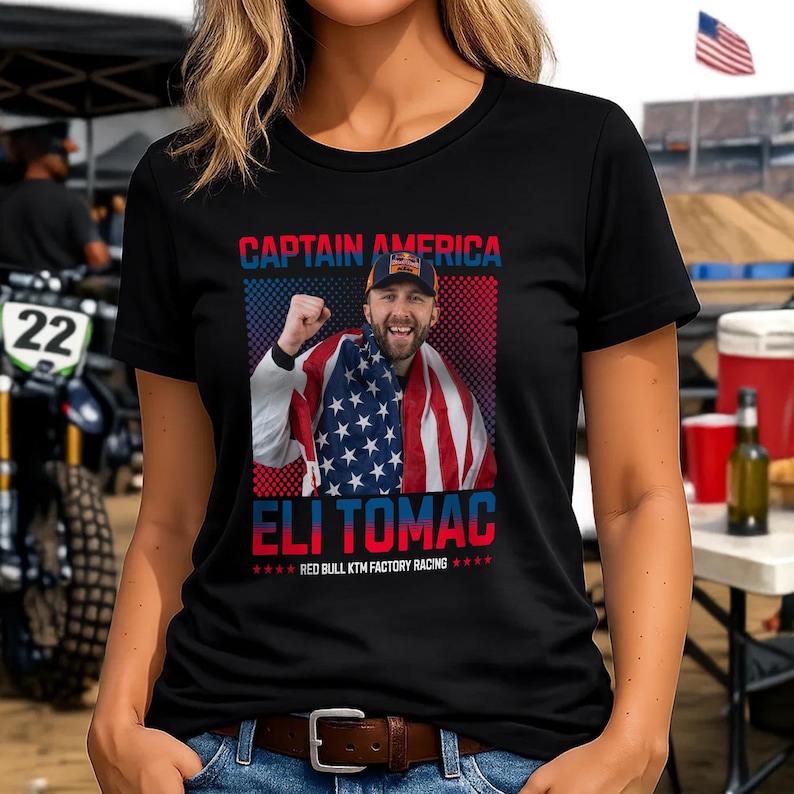 Peut inclure: T-shirt noir &agrave; col rond avec un graphisme d'un homme envelopp&eacute; dans un drapeau am&eacute;ricain. Le texte "CAPTAIN AMERICA" et "ELI TOMAC" sont affich&eacute;s au-dessus et en dessous de l'image, avec "RED BULL KTM FACTORY RACING" en dessous.