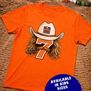 May include: Orange t-shirt featuring a graphic of a cowboy hat with the Red Bull KTM Racing Team logo, long hair, a mustache, and the number 7. The shirt has the text "AVAILABLE IN KIDS SIZES" in a blue circle.