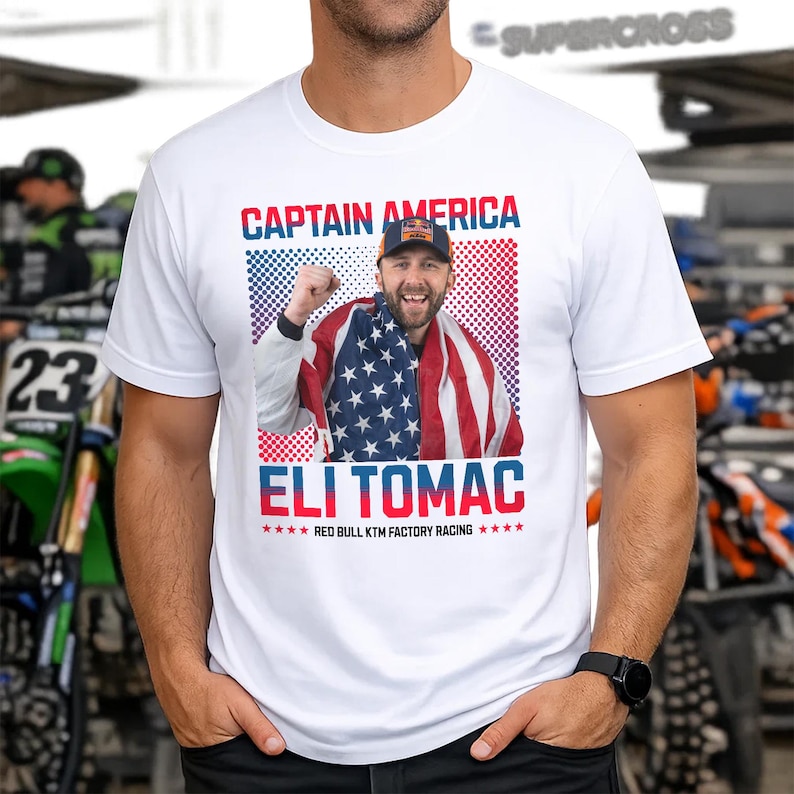 Peut inclure: T-shirt blanc avec un graphisme d'Eli Tomac, avec le texte "CAPTAIN AMERICA" et "ELI TOMAC". Le design comprend une photo de Tomac envelopp&eacute; dans un drapeau am&eacute;ricain, avec le logo Red Bull KTM Factory Racing.