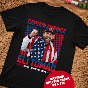 Peut inclure: T-shirt noir avec un graphisme d'Eli Tomac, envelopp&eacute; dans un drapeau am&eacute;ricain, avec le texte "CAPTAIN AMERICA" et "ELI TOMAC". Le t-shirt comprend &eacute;galement "RED BULL KTM FACTORY RACING" et "DAYTONA CHIPPED TOOTH USA TEE".