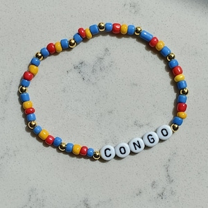 May include: A colorful beaded bracelet with blue, yellow, and red beads and gold spacers. The bracelet spells out the word "CONGO" in white letters.
