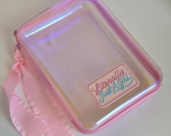 May include: A rectangular, iridescent clear pouch with a pink zipper and trim. A pink fabric patch is stitched to the front, featuring the embroidered text "Literally Just A Girl" in pink and blue script. A pink ribbon is tied to the zipper pull.