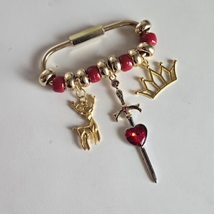 May include: A gold-toned carabiner keychain featuring alternating red and metallic gold beads. Three charms hang from the carabiner: a gold-toned deer, a silver-toned sword with a red heart-shaped gemstone, and a gold-toned crown outline.