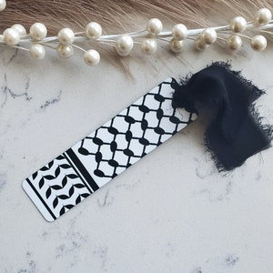 May include: A black and white bookmark with a traditional Palestinian keffiyeh pattern. The bookmark has a black ribbon attached to it.