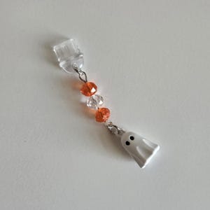 May include: A Halloween-themed charm featuring a small white ghost with black eyes, connected by a silver chain to orange and clear beads. The charm is attached to a clear plastic piece.