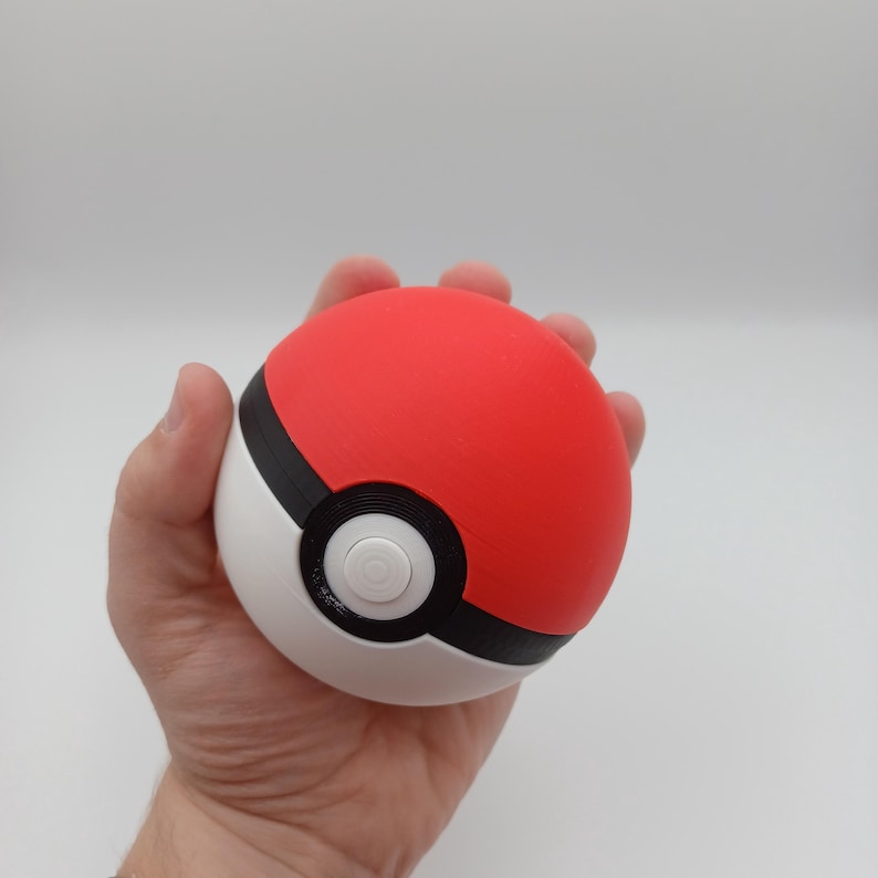 May include: A red, white, and black Pok&eacute; Ball. The top half is red, the bottom half is white, and a black band separates the two. A white button is in the center. The ball is held in a hand.