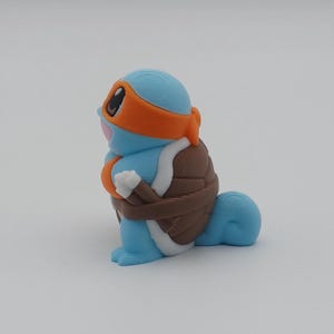 May include: A blue and brown 3D printed figurine of a turtle wearing an orange bandana. The turtle has a white underbelly and a brown shell.