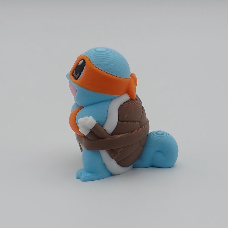 May include: A blue and brown 3D printed figurine of a turtle wearing an orange bandana. The turtle has a white underbelly and a brown shell.