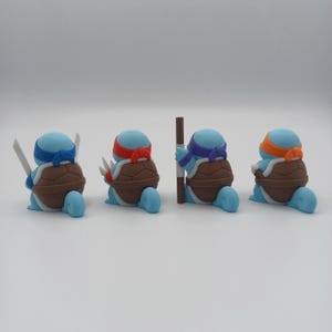 May include: Four blue turtle figurines with different coloured bandanas and weapons. The turtles are in a row, facing the right side of the image. The turtle on the left is holding two swords, the next turtle is holding two katanas, the third turtle is holding a staff, and the last turtle is holding a sai.