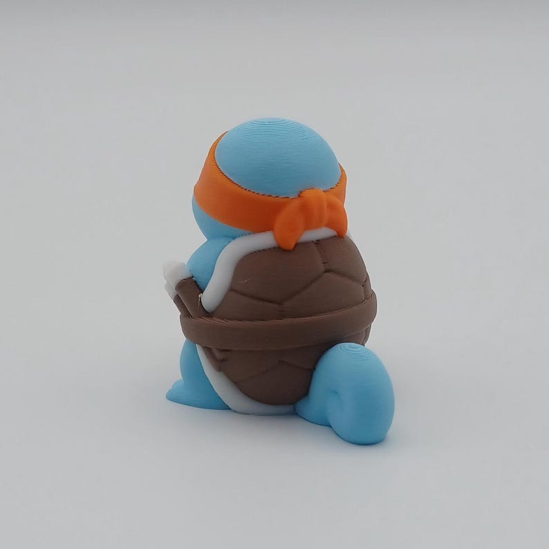 May include: A blue and brown turtle figurine with an orange bandana. The turtle is sitting on a white surface.