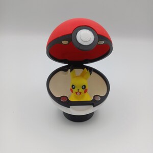 May include: A red and white Pok&eacute; Ball with a black rim, open to reveal a yellow Pikachu figure. The toy has a black base and a white button on top. The Pikachu figure has black eyes, red cheeks, and a smiling mouth.