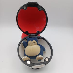 May include: A 3D-printed Pok&eacute; Ball, open to reveal a blue and tan Snorlax figure. The ball is red inside and white and black on the outside. The Snorlax is in a sleeping pose.