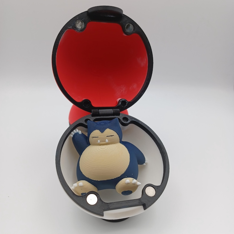 May include: A 3D-printed Pok&eacute; Ball, open to reveal a blue and tan Snorlax figure. The ball is red inside and white and black on the outside. The Snorlax is in a sleeping pose.