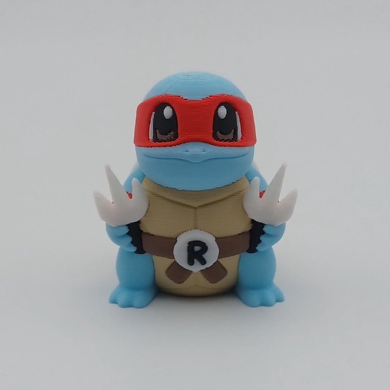 May include: A blue and white 3D printed Squirtle figurine wearing a red bandana with a black 'R' on its chest.