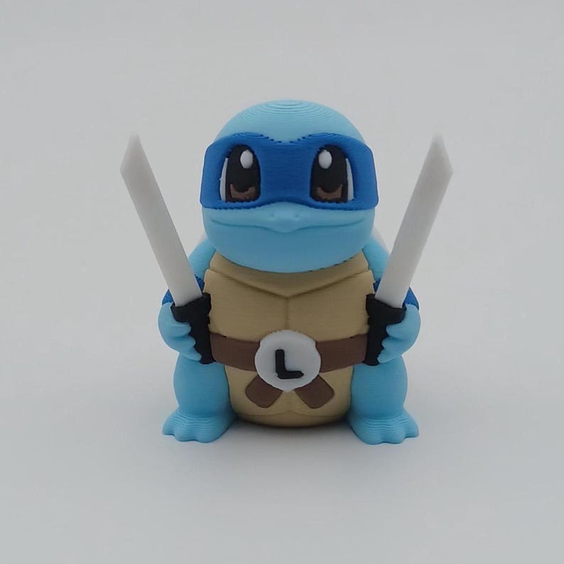 May include: A blue and brown 3D printed figurine of a turtle wearing a blue bandana and holding two white swords. The turtle has a brown belt with a white "L" on it.