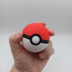 May include: A red and white Pok&eacute; Ball with a black stripe and a white button in the center. The ball is held in a hand against a white background. The ball is a sphere.